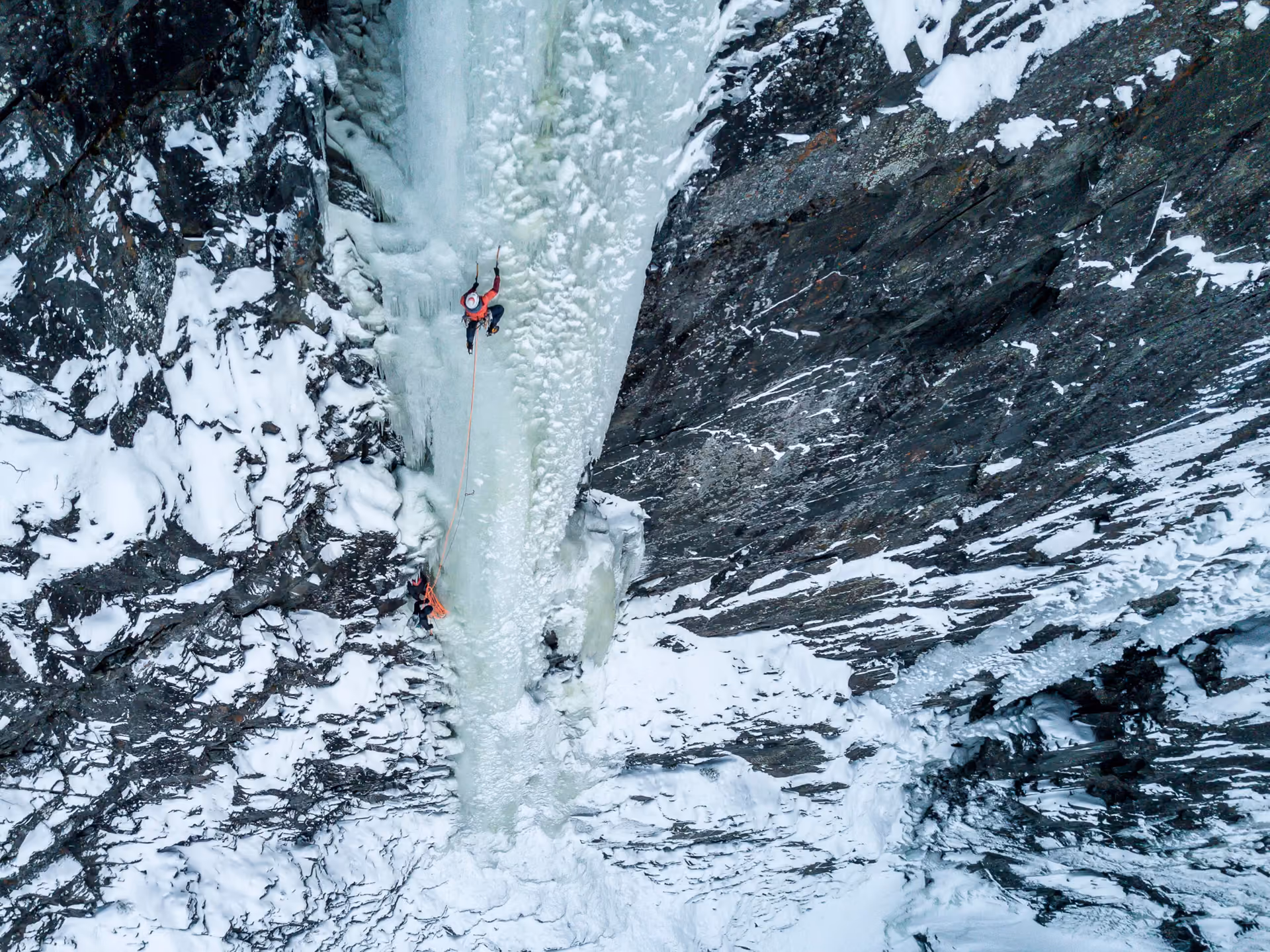 Aerial view of two climbers on a frozen waterfall between steep, snow-covered rock walls in the Juvsøyla area